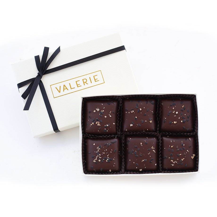 A luxurious white gift box with a black satin ribbon, containing six pieces of black sesame toffee coated in bittersweet chocolate and sprinkled with soy salt.