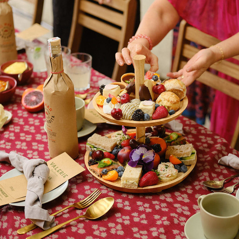 Two-tiered wooden platter with assorted finger foods on a table with wine and tea.