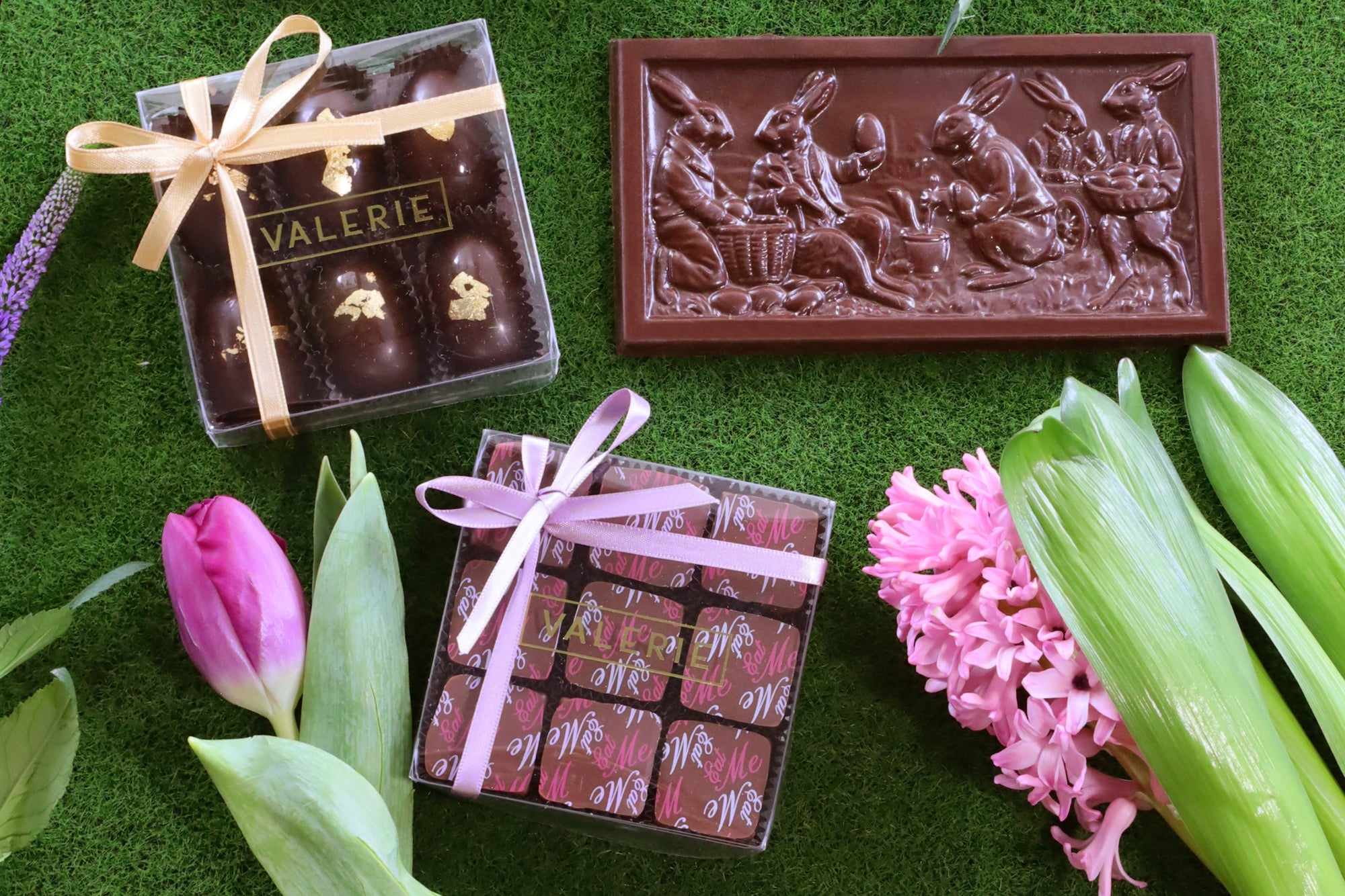 A beautifully arranged Easter chocolate collection from Valerie Confections displayed on a lush green surface. The assortment includes a gold-accented chocolate egg box tied with a gold ribbon, a festive chocolate bar embossed with an illustration of rabbits, and a spring-themed truffle box tied with a lavender ribbon. Fresh tulips, hyacinths, and greenery add a vibrant seasonal touch.