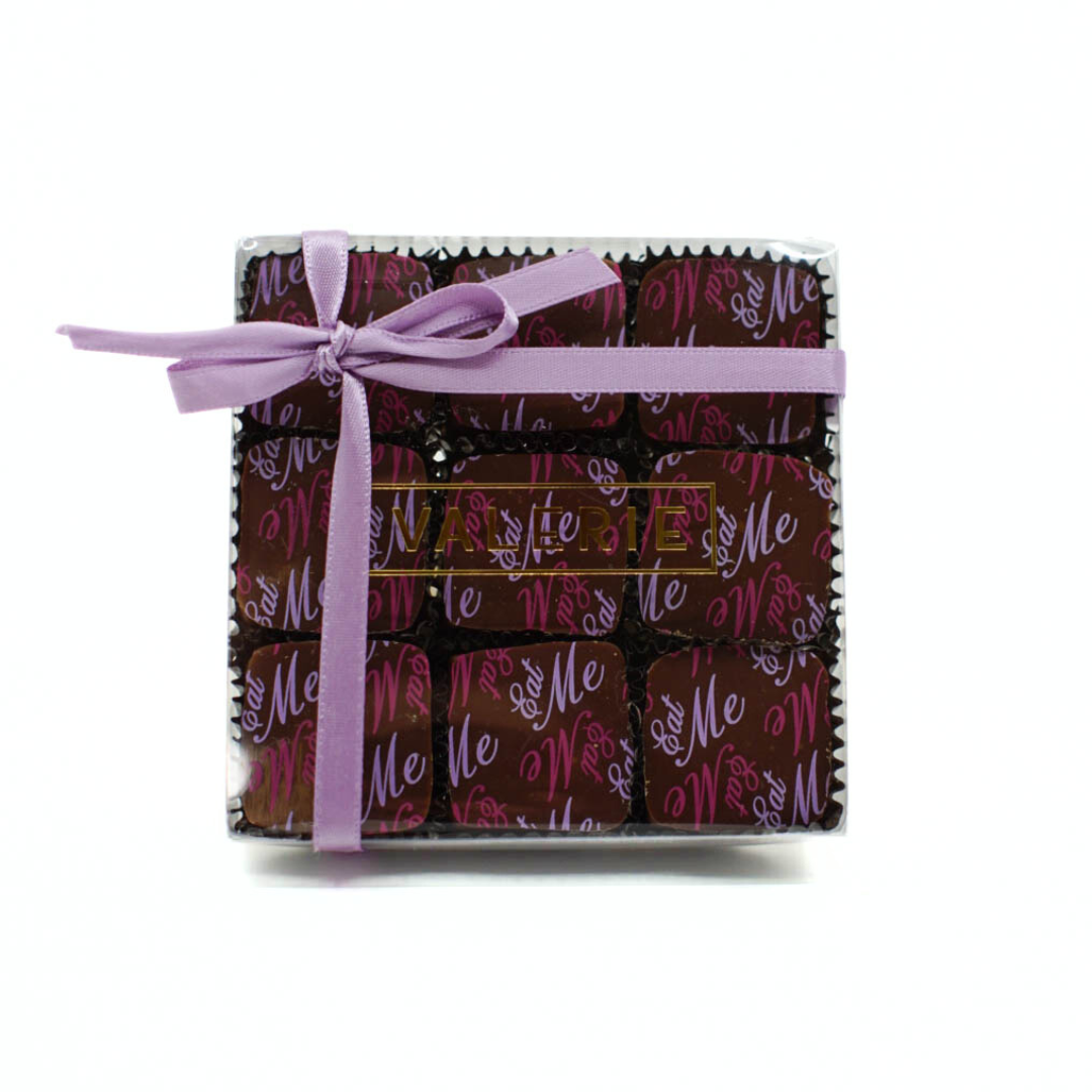 A clear box of Earl Grey-infused chocolate truffles, wrapped in darkened milk chocolate and decorated with a whimsical “Eat Me” design, tied with a plum satin ribbon.