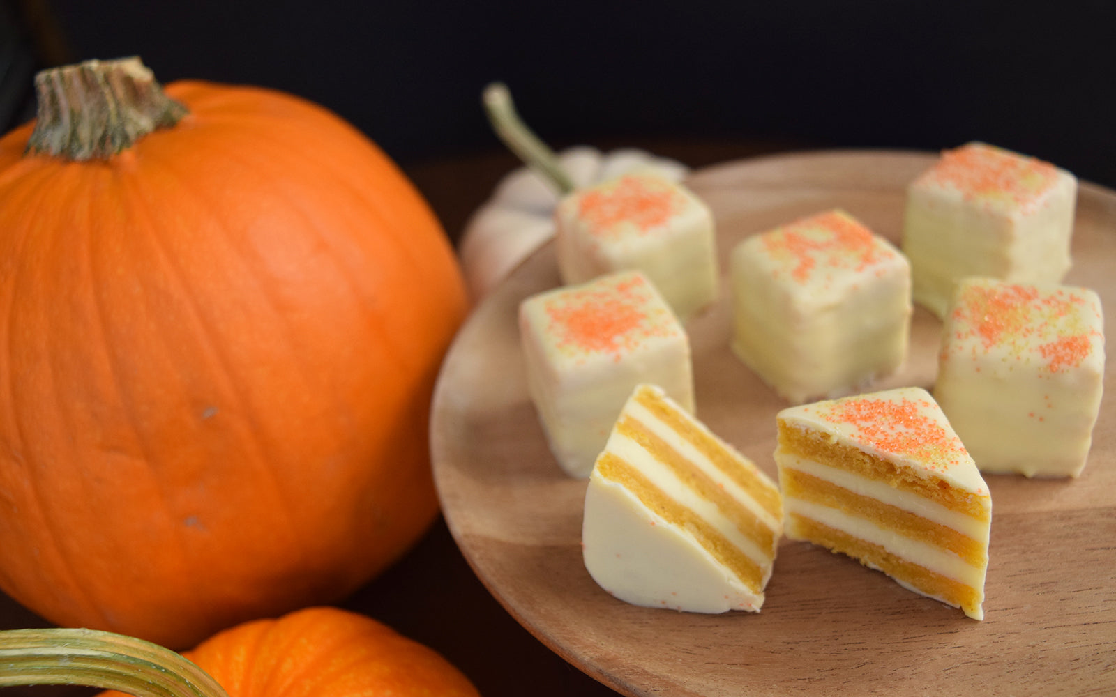 Pictured are Pumpkin Petits Fours on a wooden tray, sliced to highlight the delicate layers, with a pumpkin in the background. 