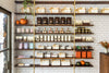 Chocolate Boxes, chocolate bars, hot chocolate mix, and other sweets displayed on walnut and brass shelves, against a subway tile wall.