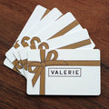 A set of elegantly designed Valerie Confections Gift Cards, featuring a gold ribbon illustration on a white background, fanned out on a wooden surface. The perfect gift for chocolate lovers.