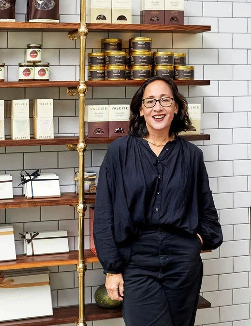 Woman in a black outfit standing in front of shelves with products on a tiled wall background