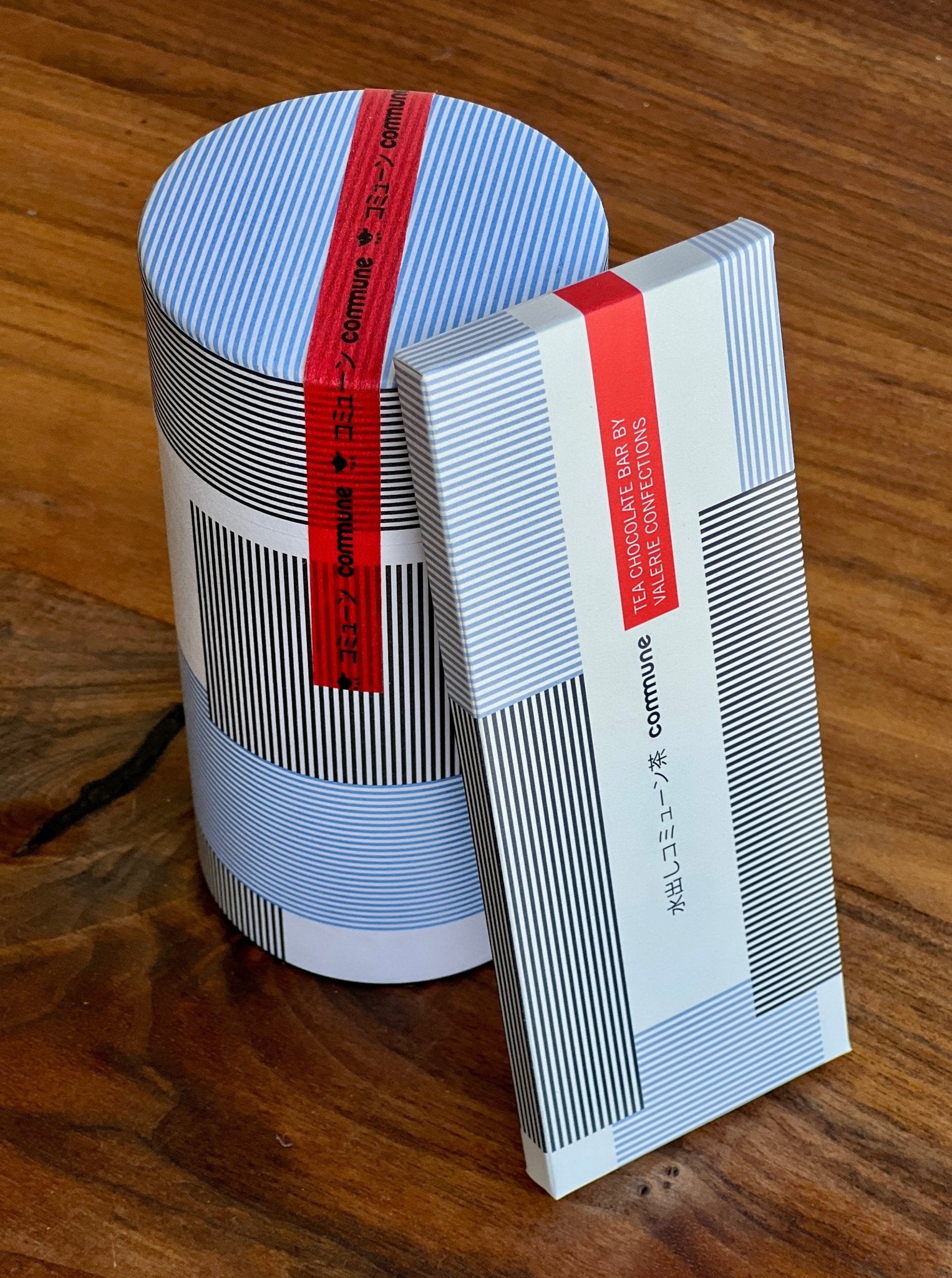 Modern-designed packaging of a cocoa product: cylindrical container and a rectangular chocolate bar, both featuring blue, white, and black minimalistic stripes and a red band with the brand name "commune".