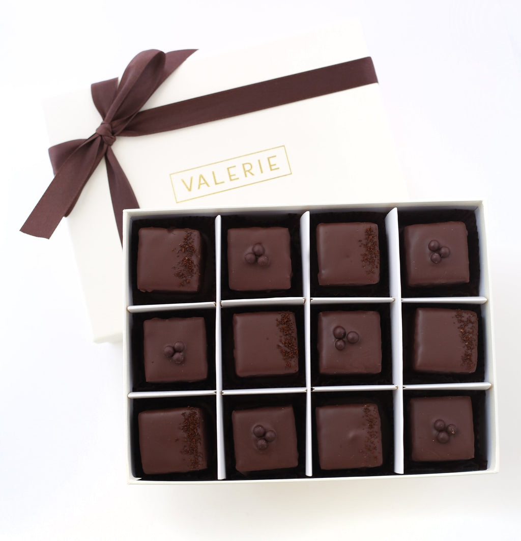 An elegant box of Valerie Confections’ Chocolate & Espresso Petits Fours, wrapped in a signature white and gold box with a chocolate satin ribbon, showcasing handcrafted gourmet treats.