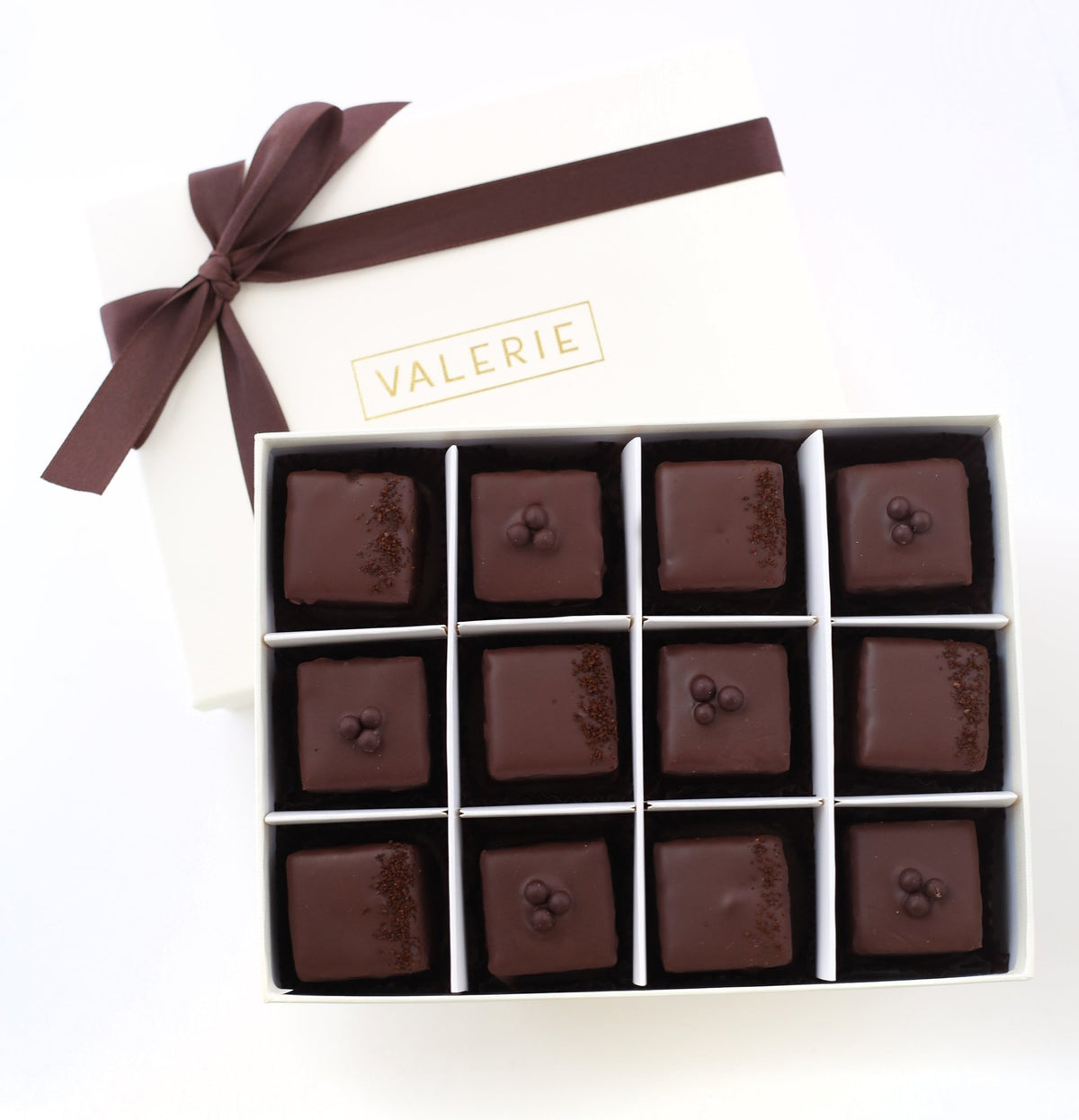 An elegant box of Valerie Confections’ Chocolate &amp; Espresso Petits Fours, wrapped in a signature white and gold box with a chocolate satin ribbon, showcasing handcrafted gourmet treats.
