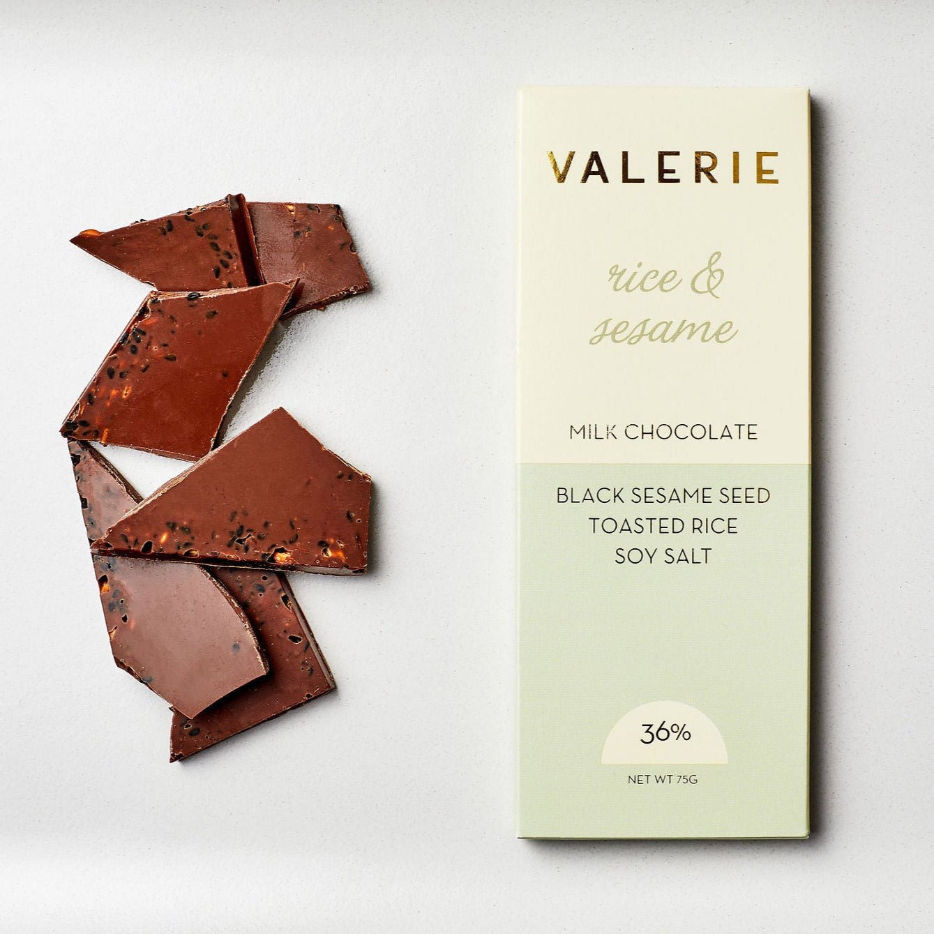 A Rice & Sesame Chocolate Bar featuring milk chocolate with black sesame seeds, toasted rice, and soy salt, elegantly packaged with broken chocolate pieces beside it.