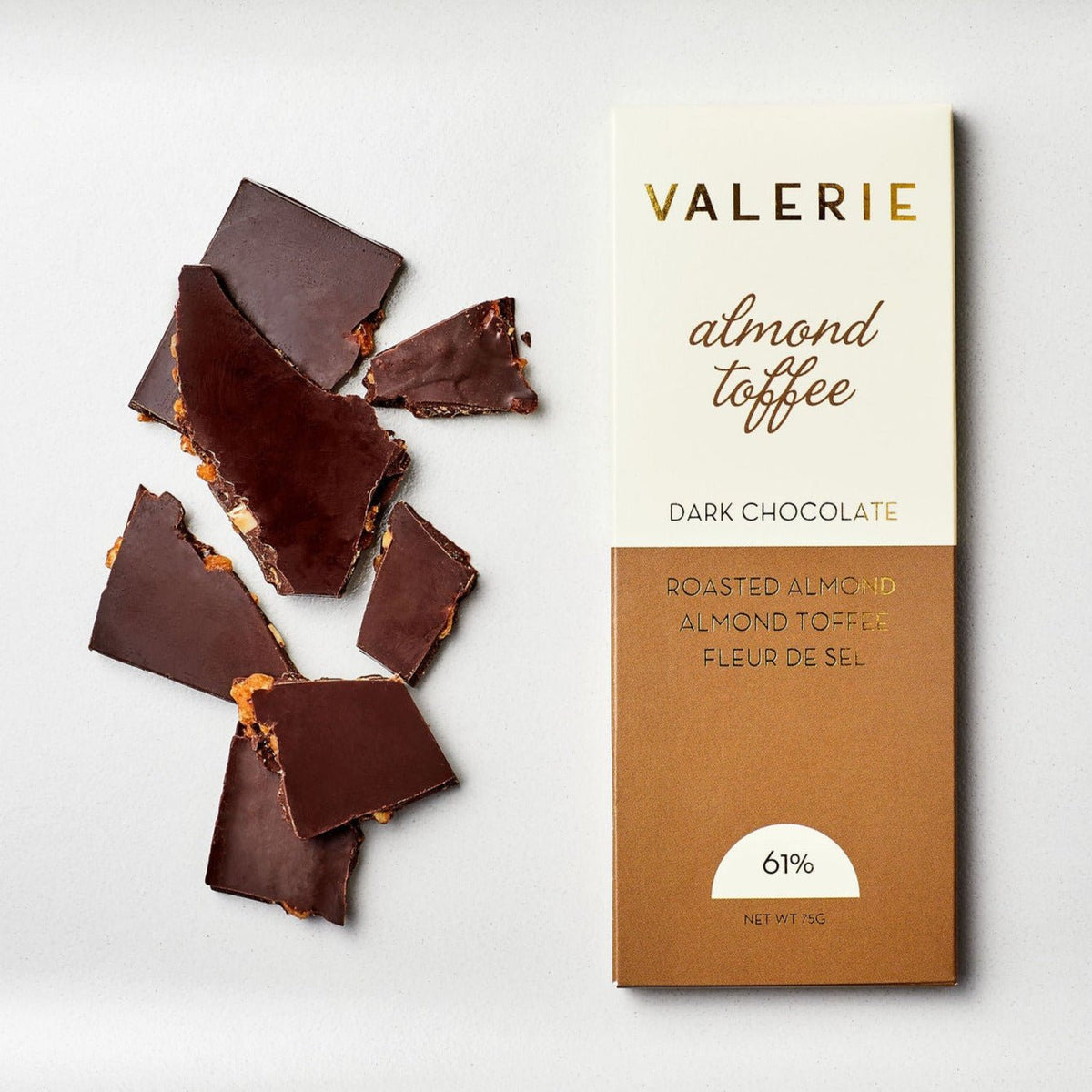 Valerie&#39;s almond toffee dark chocolate with Fleur de Sel is shown. Beside the packaging are broken pieces of the chocolate revealing almond toffee inside. Net weight is 75 grams and 61% cocoa.