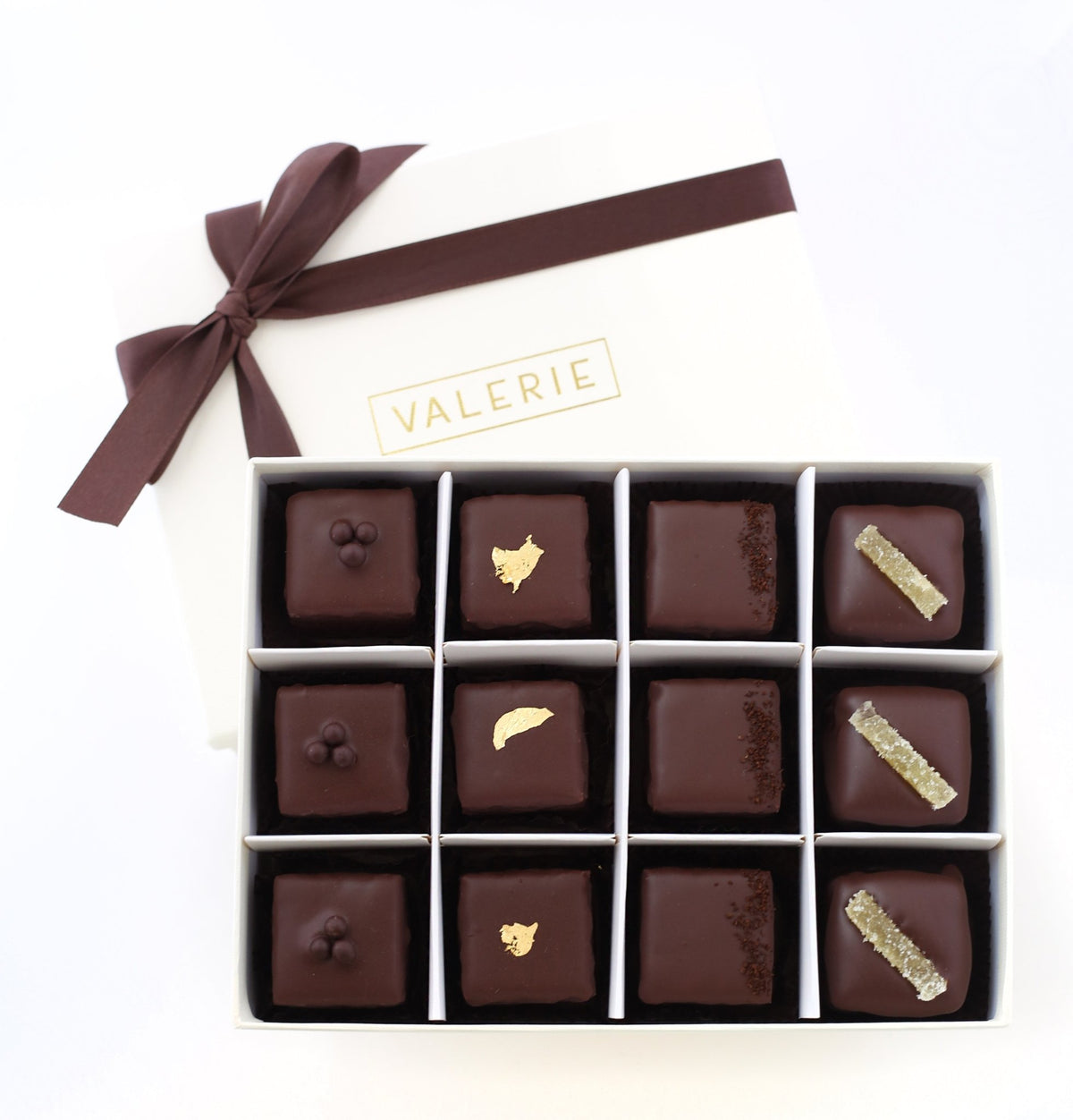 A box containing twelve chocolate-covered petits fours, some garnished with gold leaf, chocolate balls, espresso grounds, or candied ginger, with &quot;VALERIE&quot; branding on the lid.