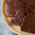 Chocolate-dipped matzo pieces sprinkled with fleur de sel on a wooden plate, resting on a gray cloth.