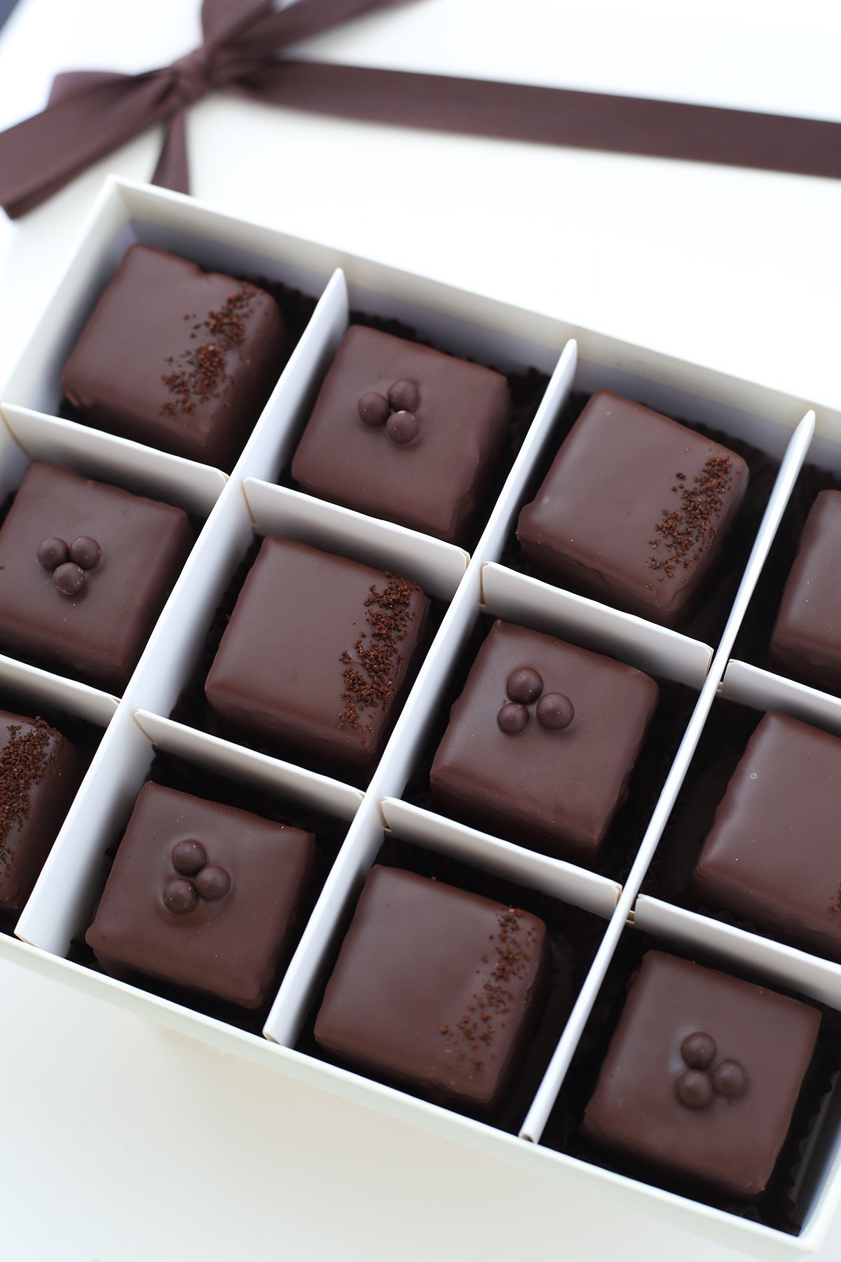 A box of 12 chocolate-covered petits fours arranged in three rows. Some are topped with chocolate beads, others with coffee grounds.