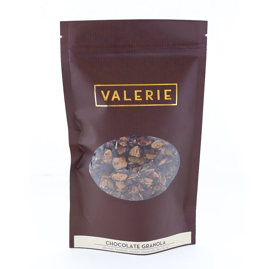 A brown pouch labeled &quot;VALERIE&quot; containing chocolate granola with visible nuts and seeds through a transparent window.