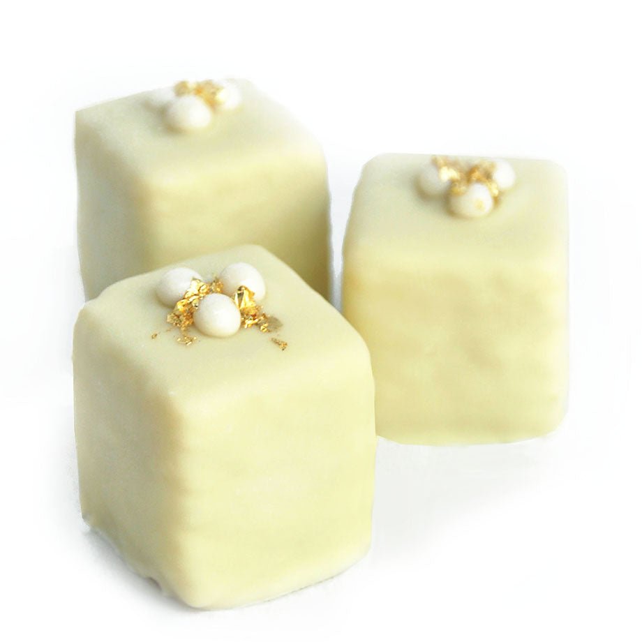 Three cream-colored eggnog petits fours decorated with pearl-like sugar decorations and gold flakes on top.