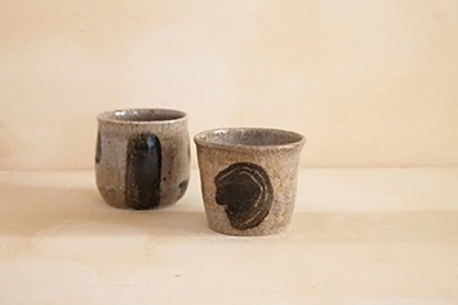 Two handcrafted gray ceramic cups with black patterns, placed on a light beige surface.