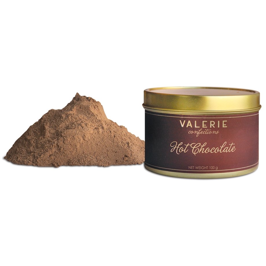 Pile of brown hot chocolate powder beside a cylindrical metal tin with a brown label that reads &quot;VALERIE Confections Hot Chocolate Net Weight 100 g.&quot;