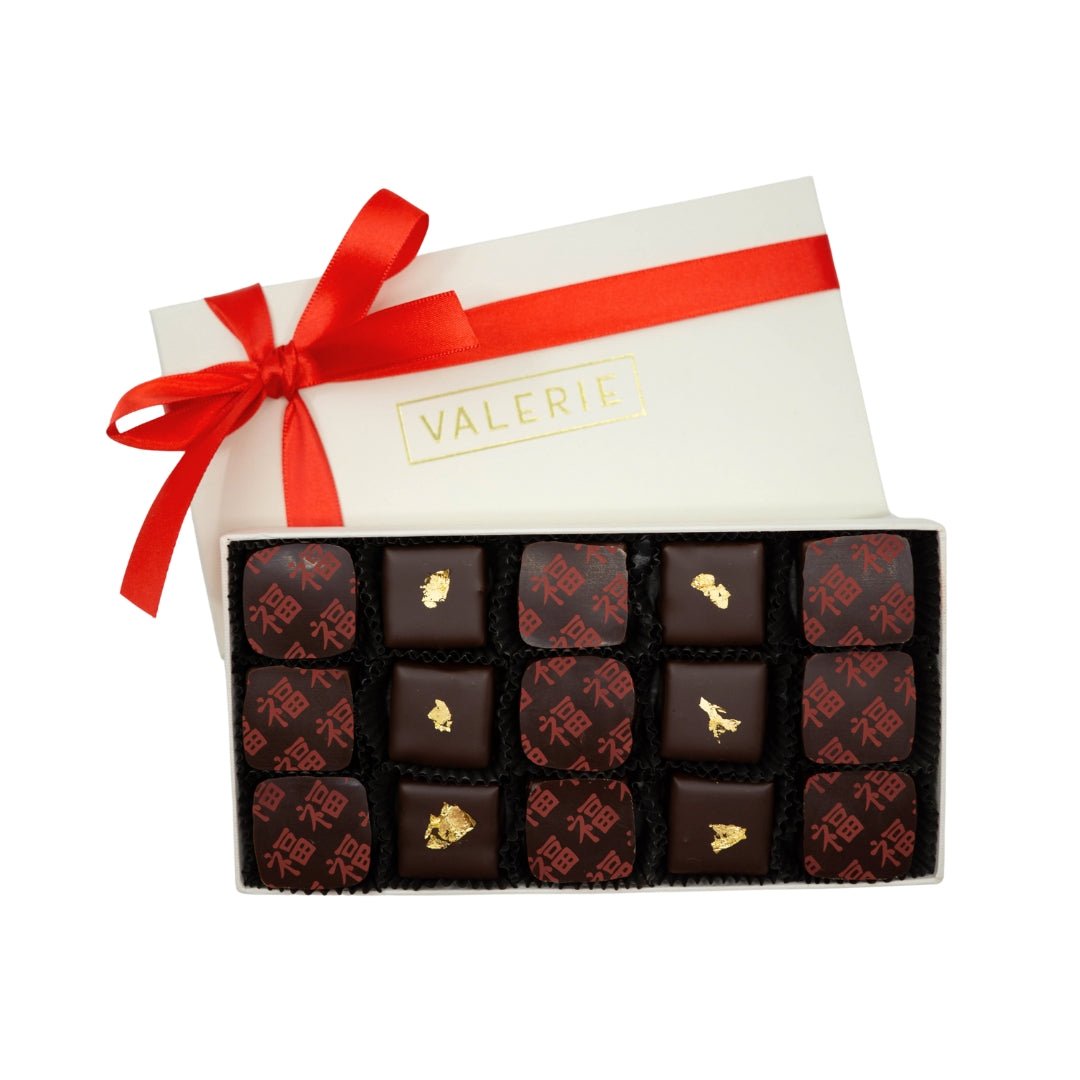 An open box of chocolates with a red ribbon and the word "VALERIE" on the lid, containing fifteen pieces with red prints and gold foil embellishments.