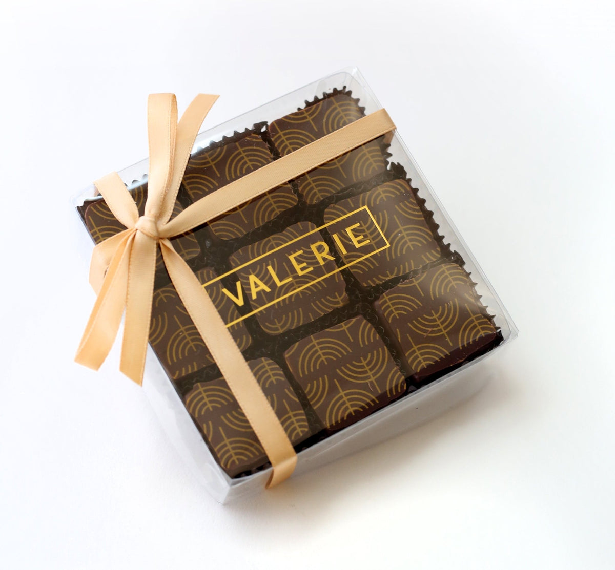 A clear plastic box with a gold satin ribbon, containing nine square dark chocolates with a gold menorah pattern. The clear box is labeled &quot;VALERIE&quot; in gold letters.