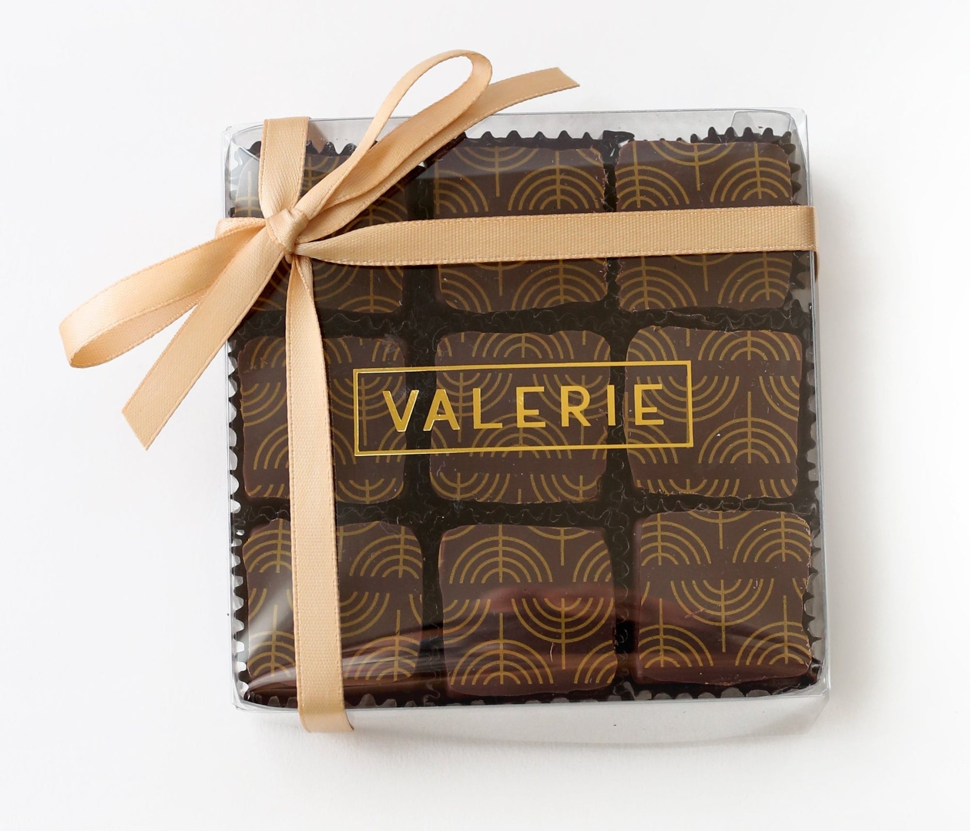 A clear plastic box with a gold satin ribbon, containing nine square dark chocolates with a gold menorah pattern. The clear box is labeled "VALERIE" in gold letters.