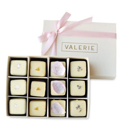 White box with pink ribbon containing twelve white chocolate petits fours with various toppings indicating different flavors.