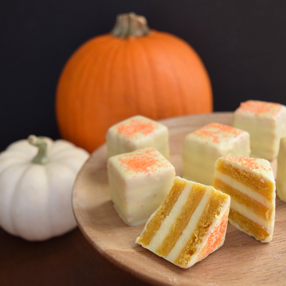 quare pumpkin petits fours with white icing and orange sprinkles on a wooden platter with a whole orange and white pumpkin in the background.