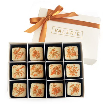 White box of twelve pumpkin petits fours with white icing and orange sprinkles, tied with an orange-brown ribbon and labeled &quot;VALERIE&quot;.