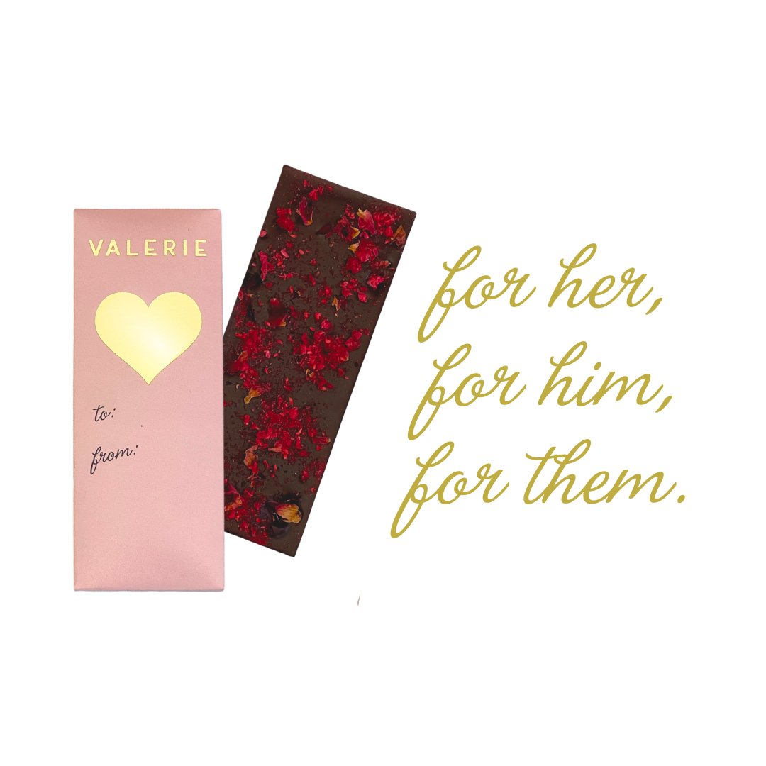 A pink chocolate bar package with a gold heart, alongside a dark chocolate bar sprinkled with red rose petals and raspberries. Text reads, &quot;for her, for him, for them.&quot;