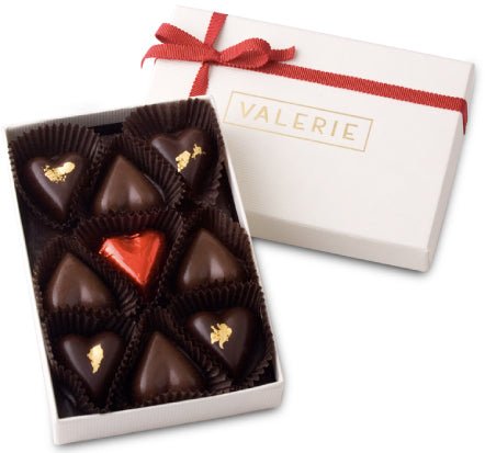 Open white box with a red ribbon containing nine heart-shaped chocolates, one wrapped in red foil, labeled &quot;VALERIE.&quot;