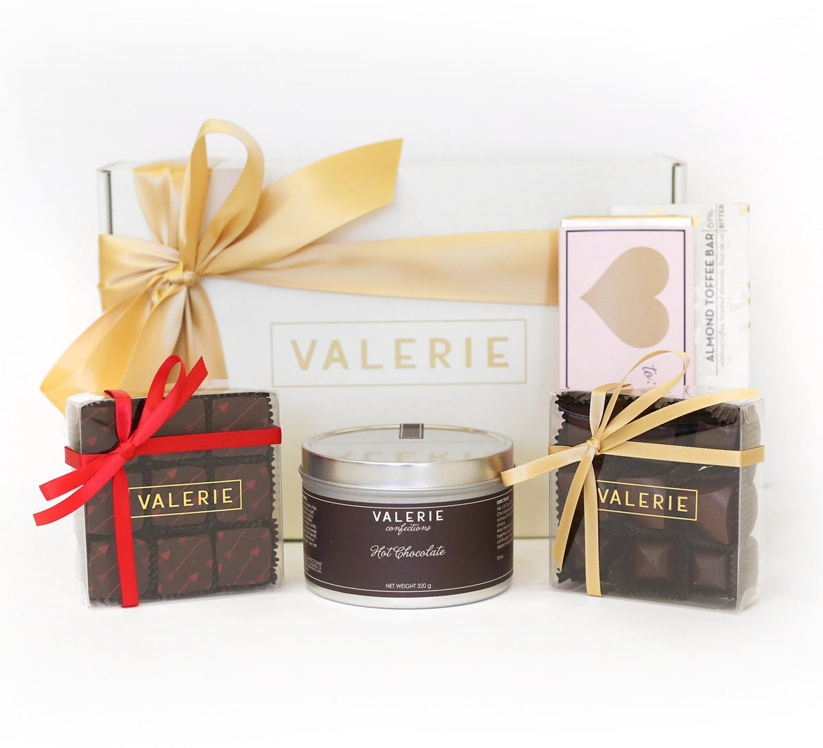 Gift box wrapped with a gold ribbon, labeled &quot;VALERIE,&quot; surrounded by a cylinder tin of hot chocolate labeled &quot;VALERIE,&quot; and two small clear boxes of chocolates with red and gold ribbons.