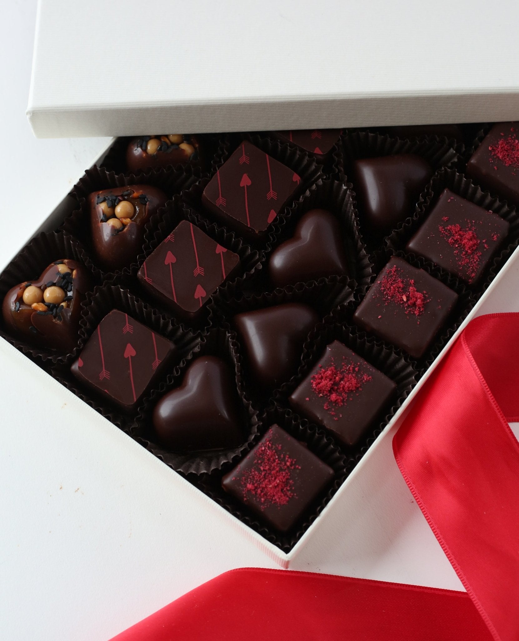 Open box of assorted chocolates with various heart shapes, on a white surface with a red ribbon.